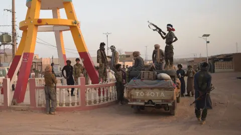 Des combattants en tenue camouflage autour d'un pick-up sur une route poussiéreuse au Mali. Un homme en silhouette, debout sur le véhicule, tient un fusil automatique levé.