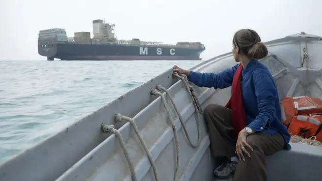 Une femme est assise dans un petit bateau gris et observe un grand cargo en mer, sur le flanc duquel sont inscrites en blanc les lettres « MSC ». Ses longs cheveux bruns sont attachés et elle porte un haut bleu. Derrière elle se trouvent quelques gilets de sauvetage orange.